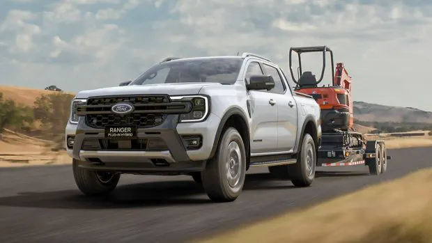 A Ford Ranger Wildtrak plug-in hybrid ute driving down a country road while towing a piece of industrial machinery